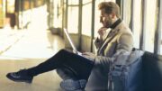 Mann im hellen Jacket checkt auf seinem Laptop Mails auf einem Flughafen und sitzt auf dem Boden mit Rucksack