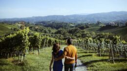 Paar mit Weinflaschen in der Hand mit dem Rücken zum Betrachter geht einen Weg in den Weinbergen der Südsteiermark