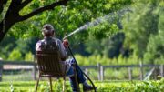 Ein alter Mann sitzt auf einem Gartenstuhl und sprengt im Sommer mit einem Schlauch den Garten.